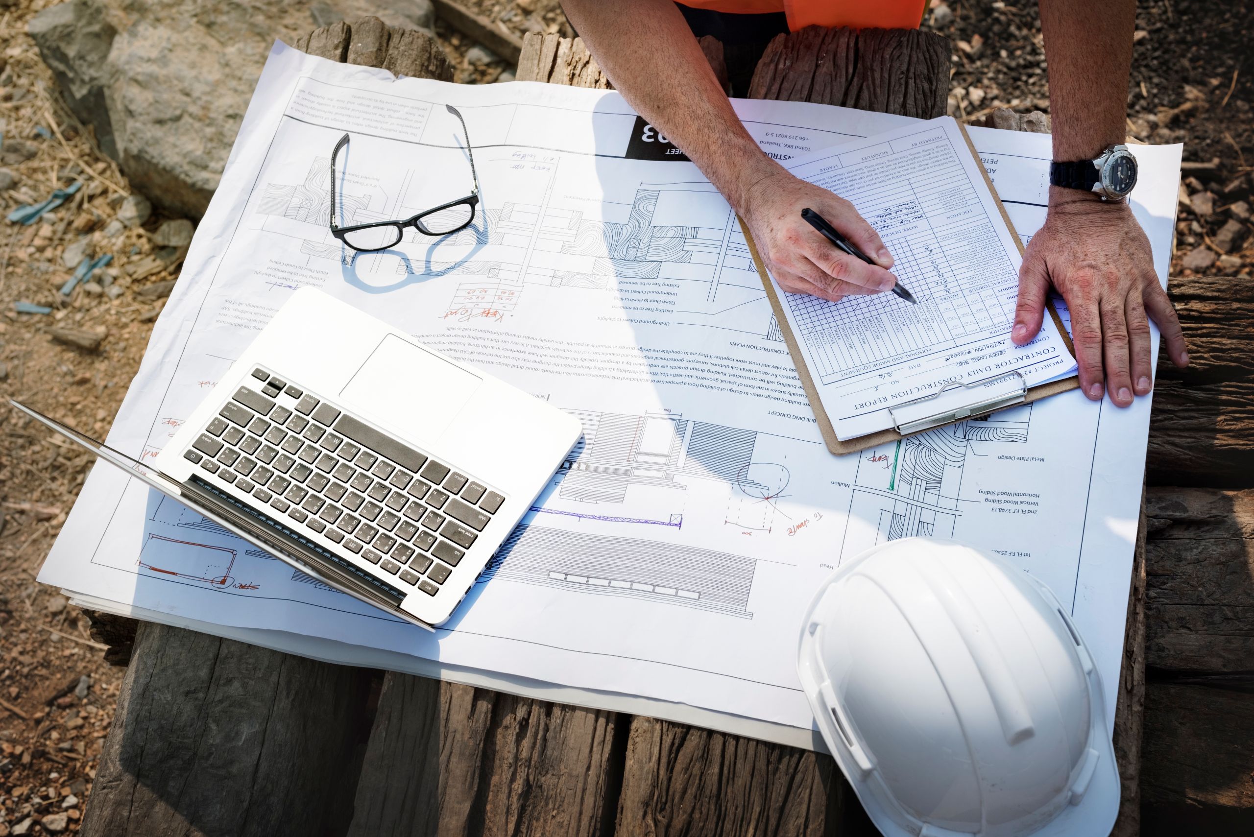 Person reviewing construction plans with laptop, clipboard, and hard hat Person reviewing construction plans with laptop, clipboard, and hard hat